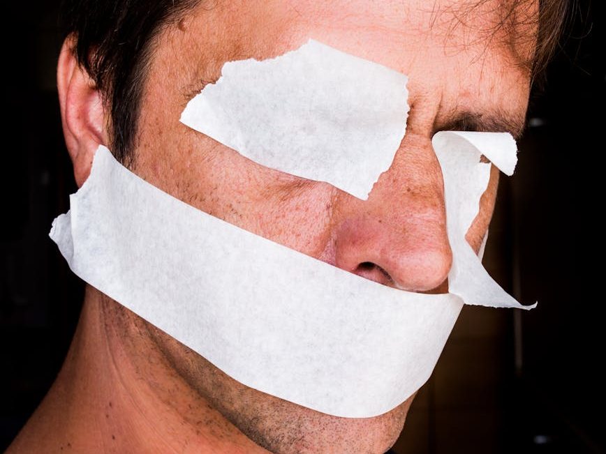 A close-up of a man's face partially obscured by white paper strips taped over his eyes and mouth, symbolizing censorship or restriction of voice.